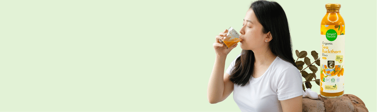 A woman in a white t-shirt drinking from a small glass cup, positioned next to a bottle of Sea Buckthorn elixir  and a small plant on a light green background.